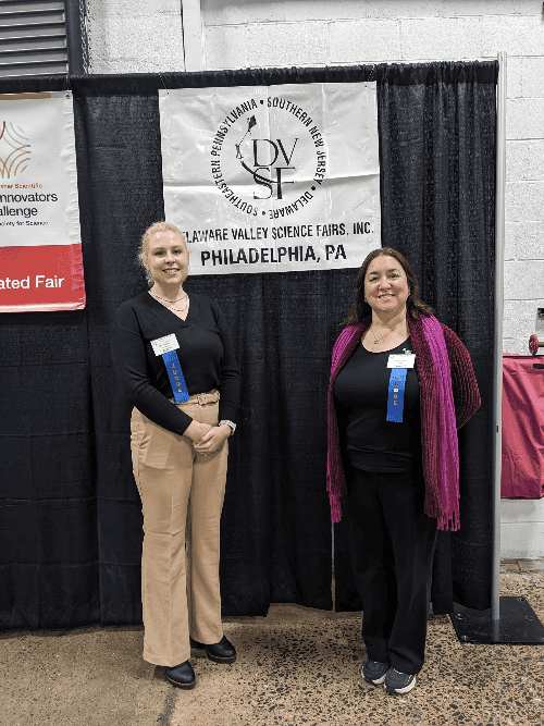 Two women with blue ribbons that indicate they are judges stand under a sign that says "Deleware Valley Science Fairs, Inc. Philadelphia, PA"