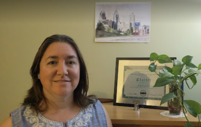 FY24 President Jen Casta smiles in a office with a green wall, framed SWE certificate and plant on a wooden cabinet, and Philadelphia skyline art on the wall.